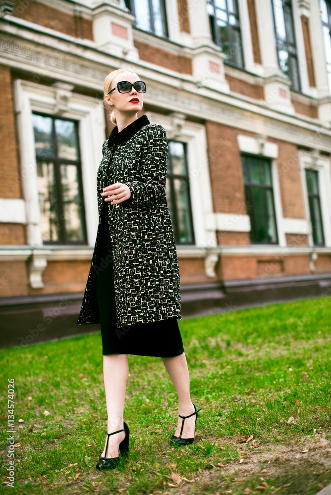 Fototapeta premium Happy Fashion blonde european elegant Woman with red lips and white skin Standing at the Old Red Brick building on green grass. Beautiful Female in formal black and white dress wearing sunglasses.