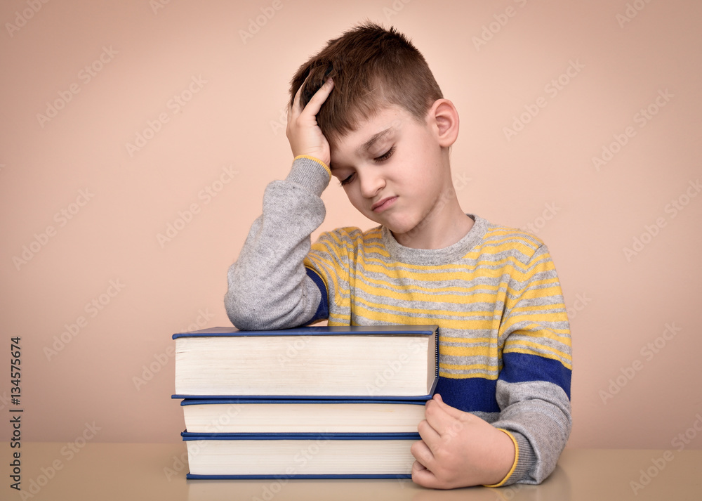 Sad and displeased young boy sitting at the table with books and holding his forehead with one hand Sad and displeased young boy sitting at the table with books and holding his forehead with one hand