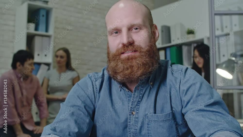 Young businessman with red beard sitting in the office and telling something at camera while his colleagues chatting in the background Young businessman with red beard sitting in the office and telling something at camera while his colleagues chatting in the background