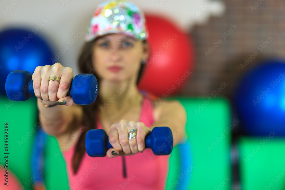 Young beautiful white girl in a pink sports suit with dumbbells in her hands before training in a fitness club. Interior of fitness club for background.