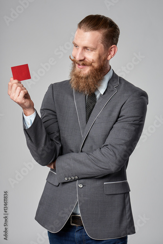 Hipster business man with beard and mustashes in suit standing over grey background holding credit card photo