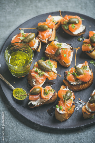 Homemade salmon crostini with cream-cheese, watercress, capers and pesto suace in round black slate stone plate over grey background, selective focus