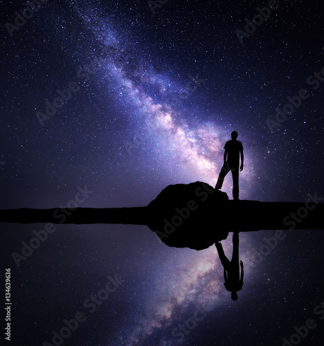 Milky Way. Night starry sky and silhouette of a standing alone man on the stone near the river with sky reflection in water. Milky way and man on the mountain. Galaxy and silhouette of a man. Universe