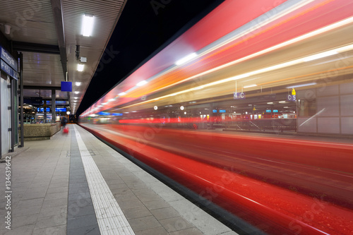 Modern railway station with high speed passenger train on railroad track in motion at night in Nuremberg, Germany. Fast blurred red commuter train.. Colorful industrial landscape