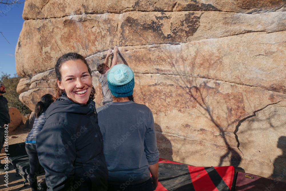 Young caucasian man climbs a granite cliff while his friends watch from below, ready to catch him if he falls. A young caucasian woman smiles at the viewer