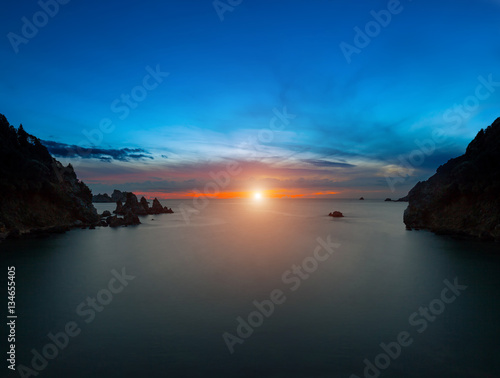 Long exposure seascape at sunset sunrise. The sun goes down  surrounded by a halo and clouds. View of the cliff into the sea and distant islands. The backlight sunbeam. Paleokastrica. Corfu. Greece.