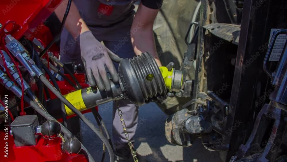 A young farmer is slowly connecting a trailor and a tractor. Then he is also putting a chain around the connector.
