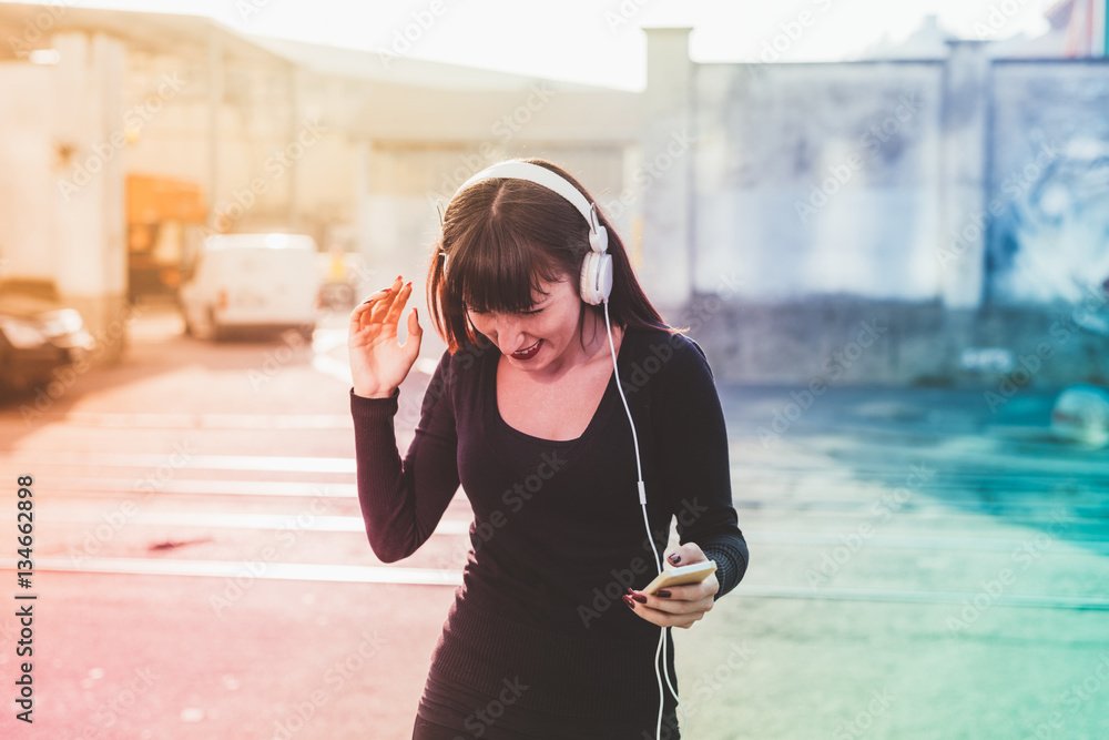 young beautiful eastern woman listening music with headphones and smart phone hand hold dancing outdoor in the city backlight, smiling - happiness, dancing, music concept
