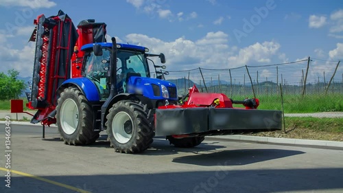 A blue tractor is standing still on a road. There is a trailor connected to it. It is a nice summer day in a village.
 photo