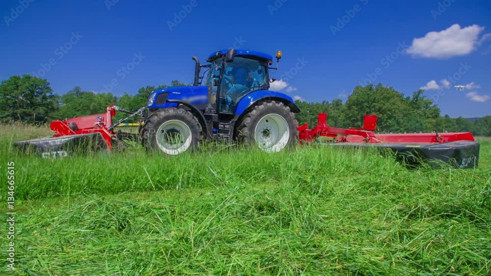 A big blue tractor is slowly driving on the field and is cutting grass with grass cutting machine. It is a gorgeous summer day.
A big blue tractor is slowly driving on the field and is cutting grass with grass cutting machine. It is a gorgeous summer day.