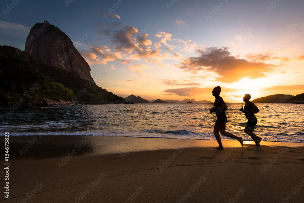Silhouettes of Two Men Running in the Beach by Sunrise with the Sugarloaf Mountain in the Horizon