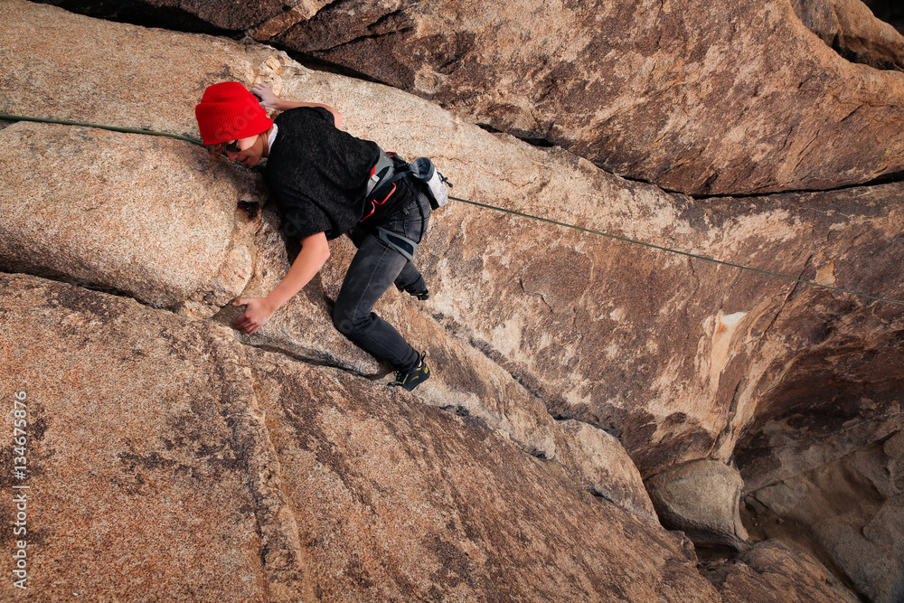 Young caucasian woman dressed for cold weather rock climbing in the desert climbs a cliff. Wearing a red hat, jeans, and sunglasses