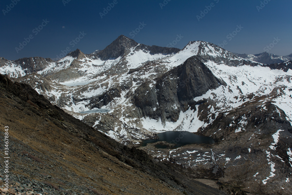Obraz premium Snow covered peaks surround a hanging lake in the high sierra in California. A hiking trail descends the foreground mountainside