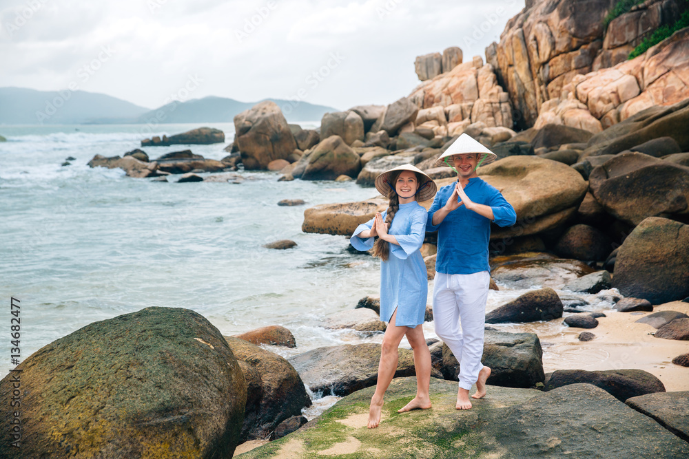 young couple in love, dressed blue shirt and Vietnamese hats in yoga pose, holding hands on the beach, looking at the sea. big stones background. Concept of family
