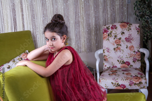 Cute lovlely middle eastern girl with dark red dress and collected hair posing and liying on green sofa at home interior. studio shot.
 photo