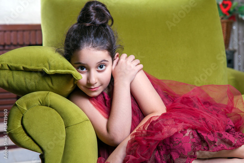 Cute lovlely middle eastern girl with dark red dress and collected hair posing and liying on green sofa at home interior. studio shot.
 photo