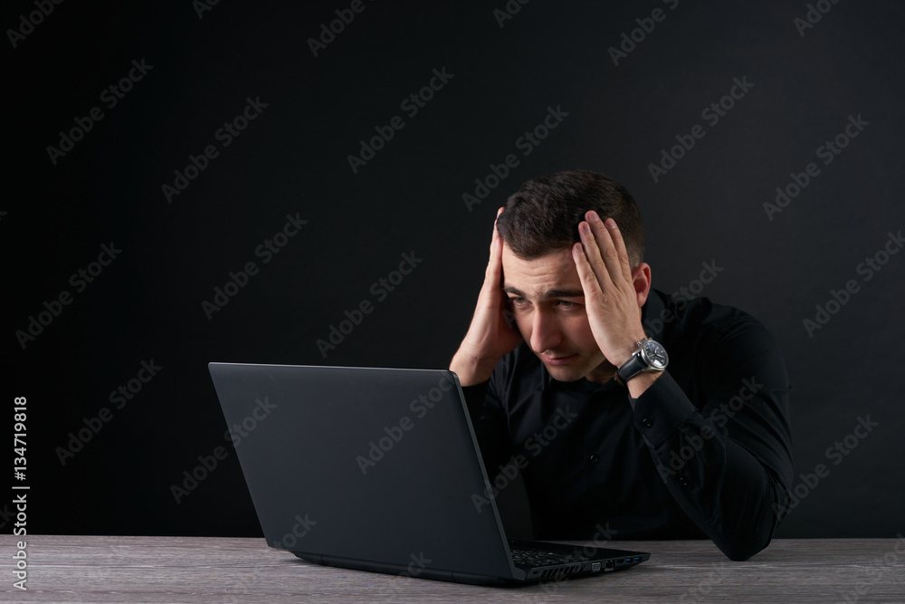 Feeling sick and tired. Frustrated young man holding head in hands while looking to laptop, Dark background with copy space Feeling sick and tired. Frustrated young man holding head in hands while looking to laptop, Dark background with copy space