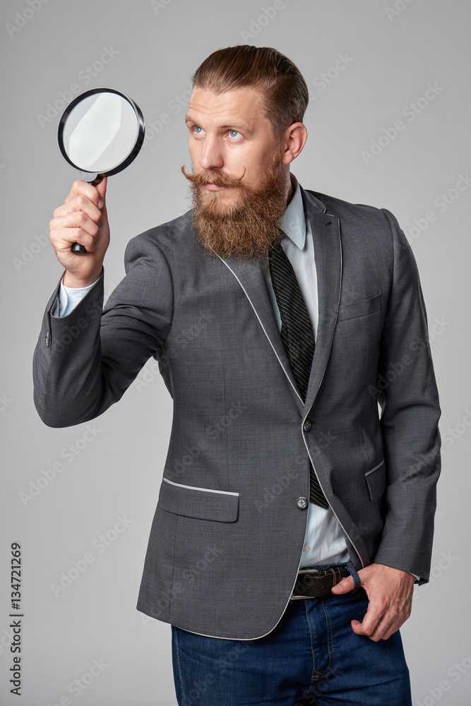 Serious hipster business man with beard and mustashes in suit looking up through magnifying glass, over grey background Serious hipster business man with beard and mustashes in suit looking up through magnifying glass, over grey background