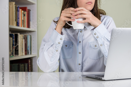 Having coffee break at white desk at home office. Girl in casual style clothes holding cup of tea or coffee sitting at laptop computer in front of bookcase