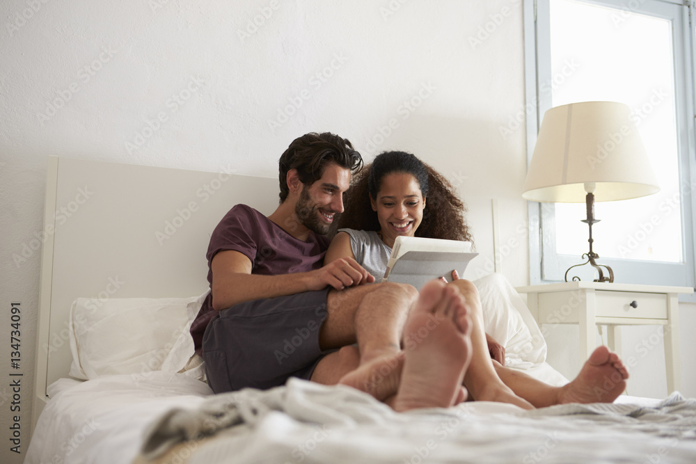 Couple Sitting On Bed Looking At Digital Tablet Together Couple Sitting On Bed Looking At Digital Tablet Together