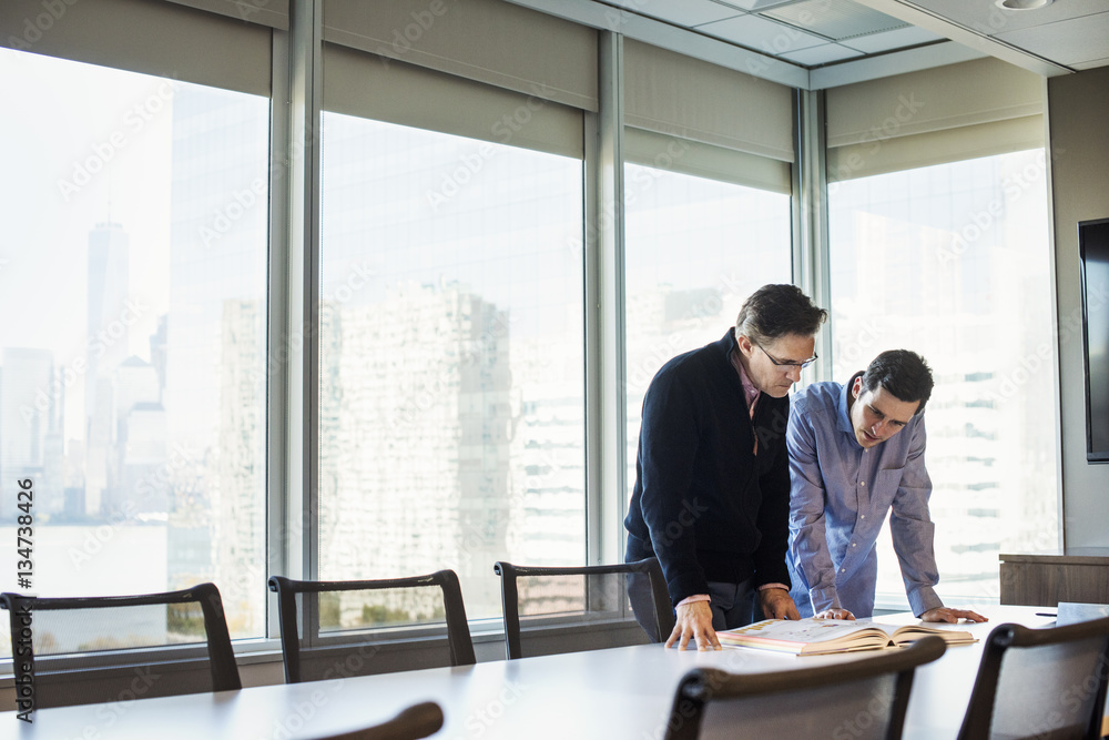 Two men standing at a table in a meeting room looking down at an open book together. Two men standing at a table in a meeting room looking down at an open book together.