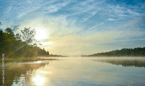 Brilliant and bright mid-summer sunrise on a lake. Warm water and cooler air at daybreak creates misty fog patches. Still water along a calm, quiet Ontario lakeside. 