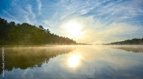 Brilliant and bright mid-summer sunrise on a lake.   Warm water and cooler air at daybreak creates misty fog patches.  Still water along a calm, quiet Ontario lakeside. 