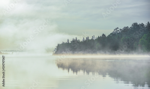 Fog and mist rises all around, partially enshrouding a waterfront deciduous Eastern Ontario forest at a lakeside. © valleyboi63