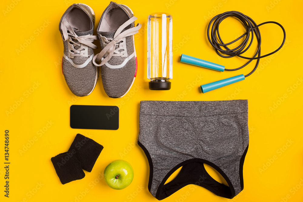 Fitness accessories on a yellow background. Sneakers, bottle of water, apple and sport top.