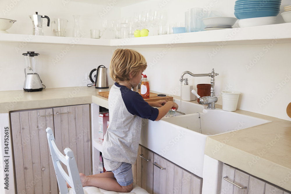 Young boy kneeling on a chair to wash dishes, full length Young boy kneeling on a chair to wash dishes, full length