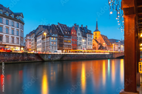 Picturesque quay and church of Saint Nicolas with mirror reflections in the river Ile and Christmas garland during evening blue hour, Strasbourg, Alsace, France