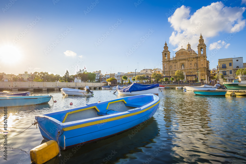 Fototapeta premium Msida, Malta - Blue traditional fishing boat with the famous Msida Parish Church at background on a summer day with blue sky and clouds