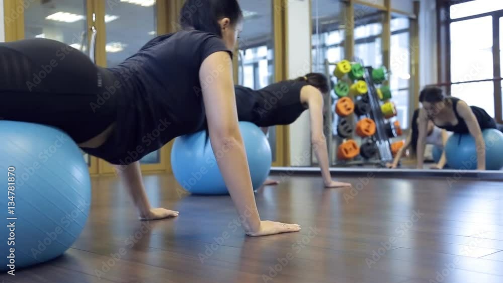 Woman athletes rock on gym ball press in fitness studio. instructor in black sportswear, showing exercises, lying on purple balloon, setting arms on floor indoors. Two fit females with picked brown