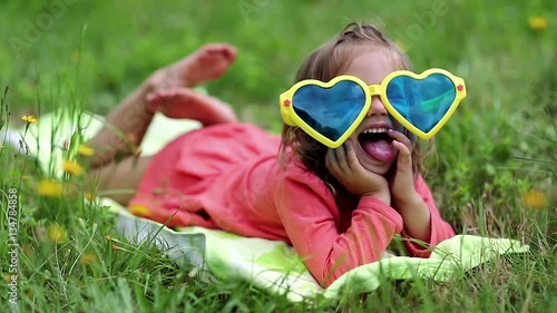 Funny little girl in glasses lies on the green grass and looks at the camera. Beautiful little girl in big sunglasses lies on the grass photo