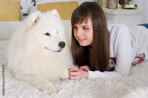 Fototapeta Naklejka Na Ścianę i Meble -  Beautiful girl lying on a fur rug and looking at the snow white Samoyed