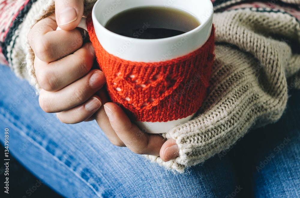 Red Knitted woolen cup with heart pattern in female hands. Hands holding a cozy Mug in red knitted mitten with hot cocoa, tea or coffee. Hands in warm concept Red Knitted woolen cup with heart pattern in female hands. Hands holding a cozy Mug in red knitted mitten with hot cocoa, tea or coffee. Hands in warm concept