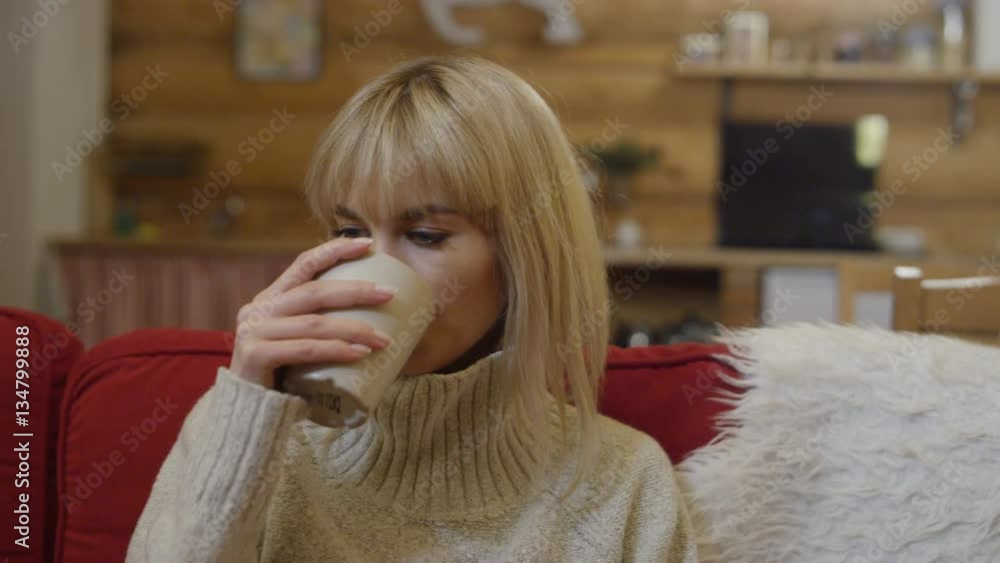 Beautiful blonde woman holding cup of tea or coffee and relaxing on a red sofa in winter wooden cabin. Shot on Red Epic. Beautiful blonde woman holding cup of tea or coffee and relaxing on a red sofa in winter wooden cabin. Shot on Red Epic.