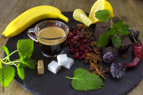 Exposition close up of fresh organic fruit and chocolate with coffe cup, half of lemon, sliced banana, mint leaf, blackberry and coffee grape on black background, rock board, healthy food.