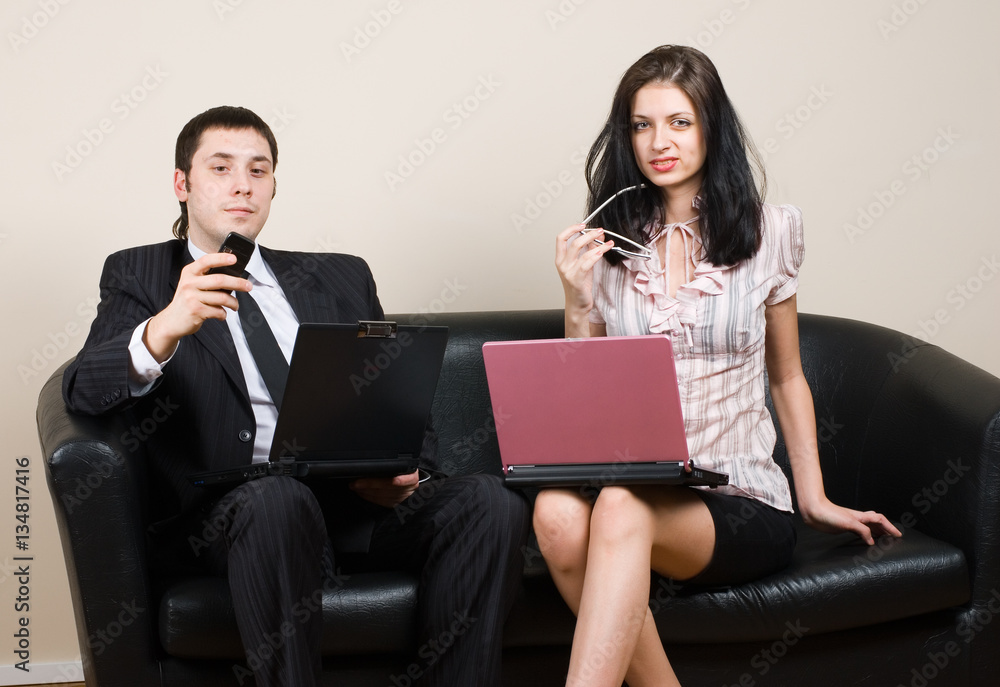 Two confident young businesspeople using a laptop and reading notes together while sitting on a sofa in a modern office Two confident young businesspeople using a laptop and reading notes together while sitting on a sofa in a modern office
