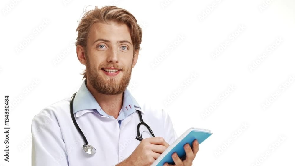 Glad Caucasian male doctor with light beard wearing medical overall smiling making notes in blue notebook on white background in slowmotion Glad Caucasian male doctor with light beard wearing medical overall smiling making notes in blue notebook on white background in slowmotion