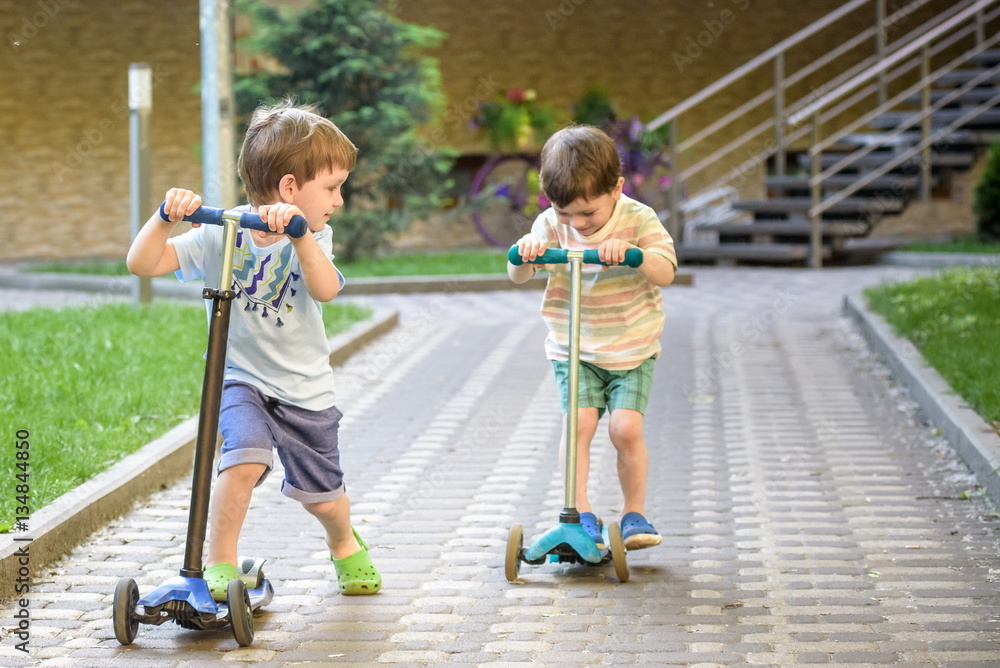 Two cute boys, compete in riding scooters, outdoor in the park,