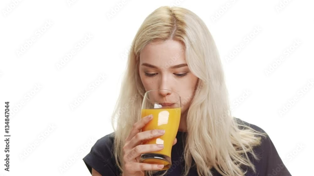 Young cute smiling blonde girl wearing dark blue shirt drinking orange juice from glass on white background in slowmotion Young cute smiling blonde girl wearing dark blue shirt drinking orange juice from glass on white background in slowmotion