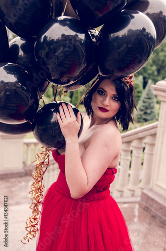 Beautiful fashion young woman in gorgeous red dress and crown holding black balloons