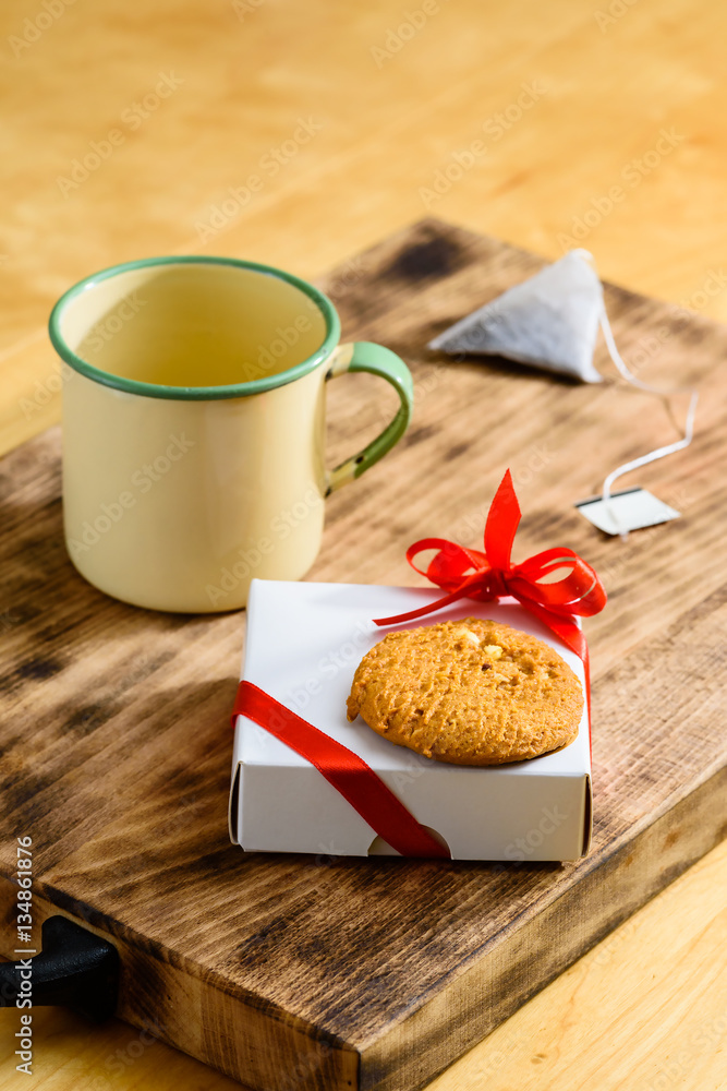 Small white present with red bow and cookie on top together with teacup and teabag on wooden tray. A humble yet lovely surprise to wake up to or find on your place at breakfast or teatime. Small white present with red bow and cookie on top together with teacup and teabag on wooden tray. A humble yet lovely surprise to wake up to or find on your place at breakfast or teatime.