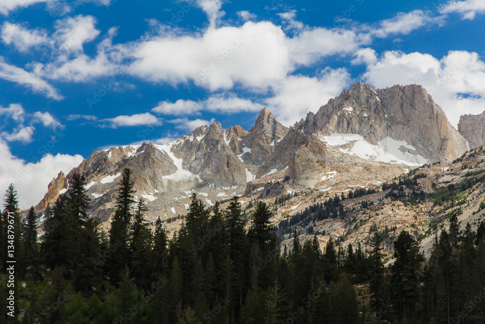 Fluffy white clouds drift over the jagged peaks of the granite mountains of California's high sierra. Below is a forested valley