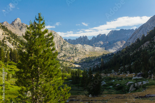Blue sky and fluffy clouds hang above jagged granite peaks over a deep forested valley in California's high sierra