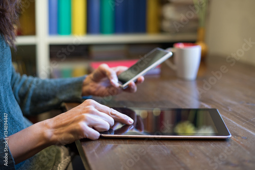 hands of adult brunette woman with green sweater touching screen tablet and using smartphone, multitasking, on wooden table with cup of coffee, library behind 