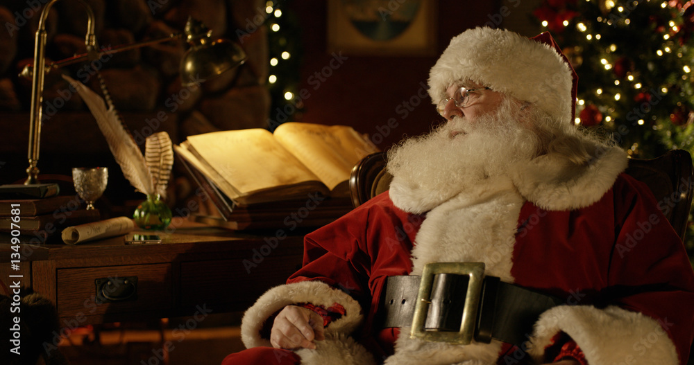 Santa Claus rests at his work bench in a room filled with Christmas decorations.