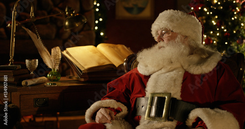 Santa Claus rests at his work bench in a room filled with Christmas decorations.