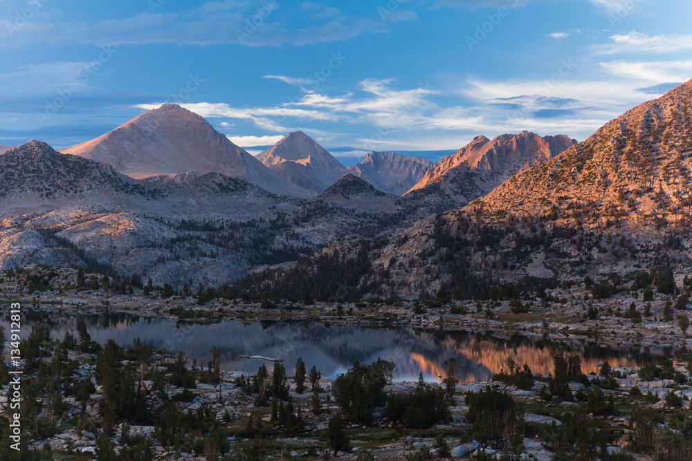 Sunset over an alpine basin with lake and pine trees surrounded by tall bare granite peaks in California